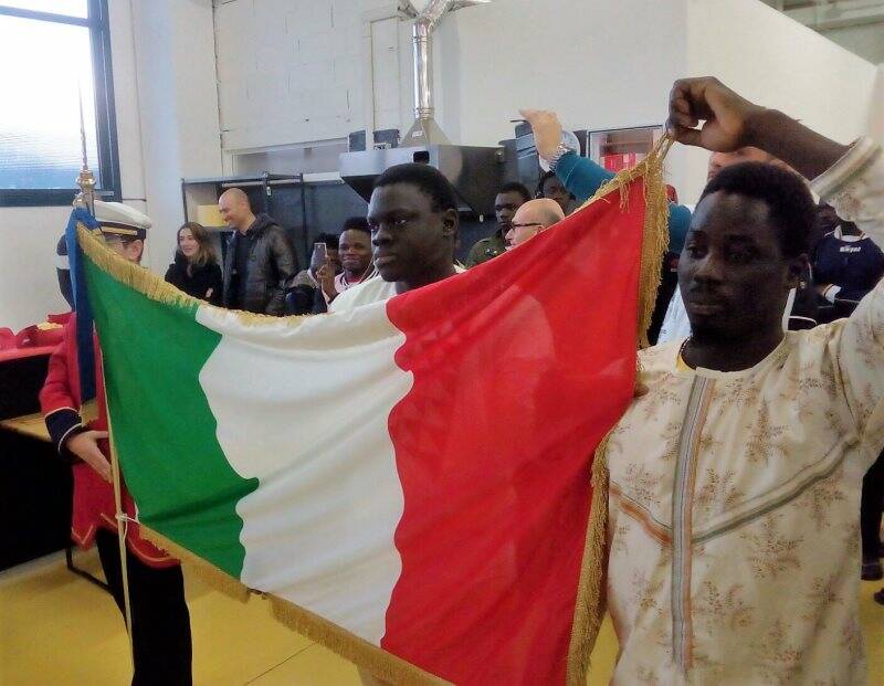 Due giovani migranti sorreggono il tricolore mentre la banda suona l'inno di Mameli